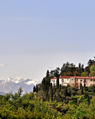 A long, pale villa with a red-tiled roof sits on a lush, tree-covered hillside, with snow-capped mountains visible in the distance across a body of water.