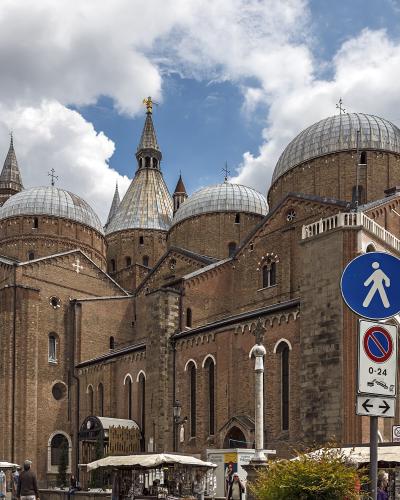 The image shows the exterior of a large brick basilica with multiple domes and spires, featuring a prominent blue pedestrian crossing sign and market stalls in the foreground under a cloudy sky.