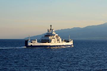 A white and blue car ferry named "RIACE" with "GLICHERNOS" branding on its side, sailing on open water with hazy mountains in the background under a pale sky.
