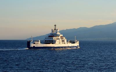A white and blue car ferry named "RIACE" with "GLICHERNOS" branding on its side, sailing on open water with hazy mountains in the background under a pale sky.