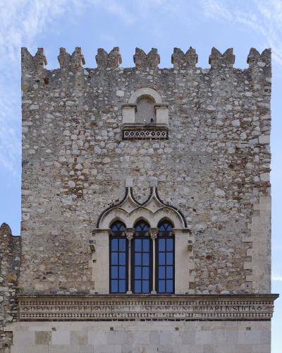 A stone medieval tower facade featuring a triple-arched Gothic window, a small balcony above it, crenellations at the top, and a Latin inscription frieze reading "DEVVM DILIGERE PRVDENTIA EST ESVM IMITARI IVSTICIA".