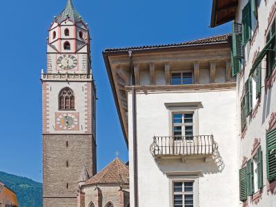 A tall, multi-tiered church tower with a clock face and a green spire stands next to a white building with green shutters in a paved town square under a clear blue sky.