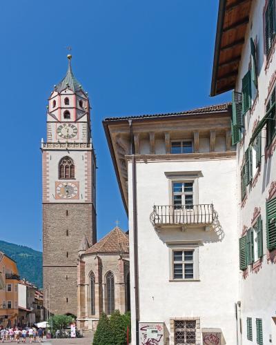 A tall, multi-tiered church tower with a clock face and a green spire stands next to a white building with green shutters in a paved town square under a clear blue sky.