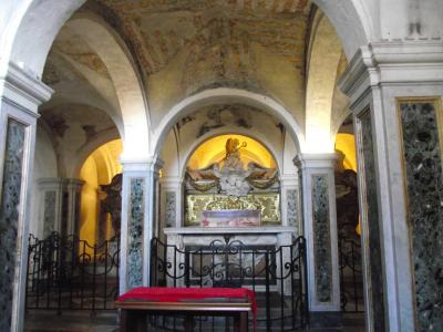 The image shows an interior view of a historic church chapel or side altar, featuring arched ceilings with exposed brick and plaster, marble-clad columns, a central stone altar with a glass reliquary, and a red cushioned kneeler in the foreground.