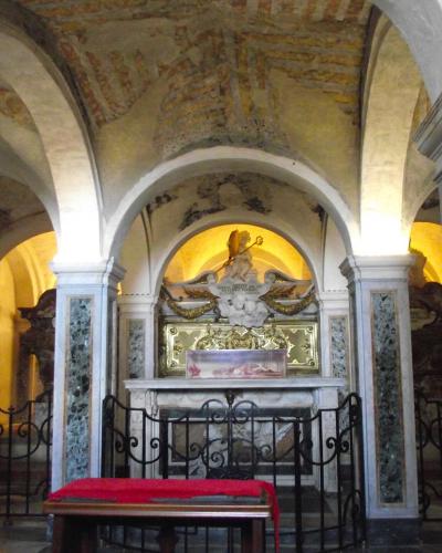 The image shows an interior view of a historic church chapel or side altar, featuring arched ceilings with exposed brick and plaster, marble-clad columns, a central stone altar with a glass reliquary, and a red cushioned kneeler in the foreground.