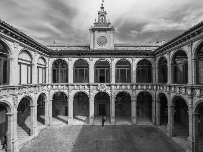 A black-and-white photograph of a two-story Renaissance-style courtyard with arched loggias on both levels, a central clock tower topped with a bell and flag, and a single person standing in the paved courtyard below.