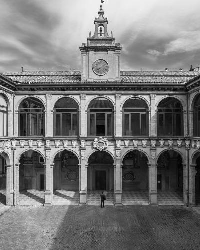 A black-and-white photograph of a two-story Renaissance-style courtyard with arched loggias on both levels, a central clock tower topped with a bell and flag, and a single person standing in the paved courtyard below.