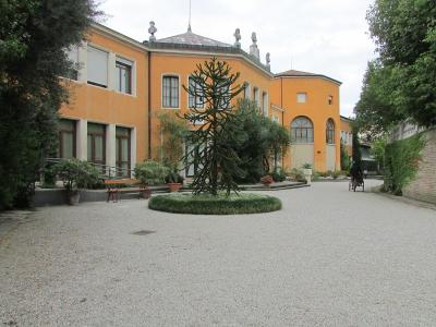 The image shows a gravel courtyard with a distinctive orange-yellow building featuring classical architectural details, statues on the roofline, and a central circular planter with a tall, spiky succulent-like plant; a person in a wheelchair is vi