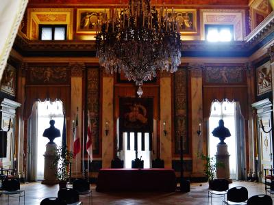 The image shows an ornate interior hall with a large crystal chandelier, marble columns, busts on pedestals, Italian and Genoan flags, and a tapestry bearing the word "GENOVA" above a central doorway.