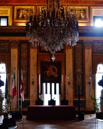 The image shows an ornate interior hall with a large crystal chandelier, marble columns, busts on pedestals, Italian and Genoan flags, and a tapestry bearing the word "GENOVA" above a central doorway.