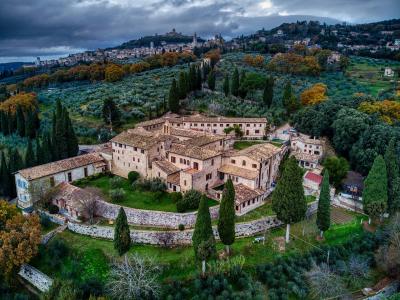 An aerial view of a large, historic stone monastery complex with terracotta roofs, surrounded by olive groves and cypress trees, with a hilltop town visible in the background under a cloudy sky.