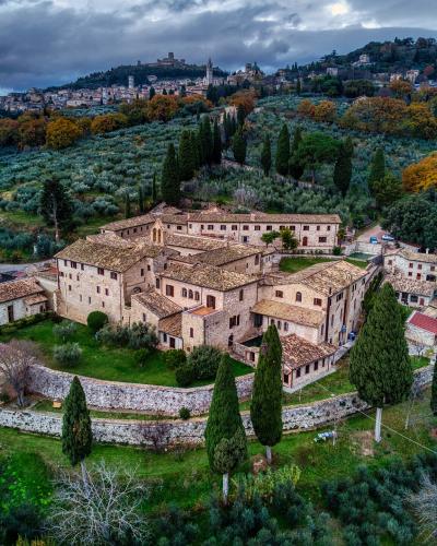 An aerial view of a large, historic stone monastery complex with terracotta roofs, surrounded by olive groves and cypress trees, with a hilltop town visible in the background under a cloudy sky.