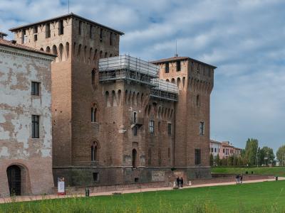 A low-angle photograph of a large, rectangular brick fortress with prominent corner towers, crenellated battlements, and arched windows, featuring modern scaffolding on one section, situated beside a grassy moat area with people walking nearby.