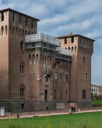 A low-angle photograph of a large, rectangular brick fortress with prominent corner towers, crenellated battlements, and arched windows, featuring modern scaffolding on one section, situated beside a grassy moat area with people walking nearby.