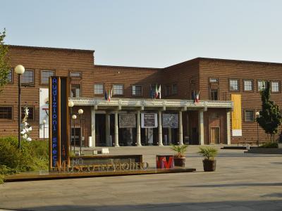 The image shows the exterior of a modern brick building with a colonnaded entrance, featuring prominent signage reading "Museo del Violino" in large metallic letters on a low platform in the foreground, along with a vertical digital sign displayin
