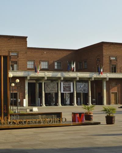 The image shows the exterior of a modern brick building with a colonnaded entrance, featuring prominent signage reading "Museo del Violino" in large metallic letters on a low platform in the foreground, along with a vertical digital sign displayin