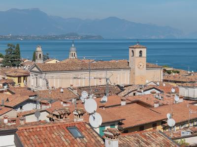A high-angle view of the terracotta rooftops of Desenzano del Garda, featuring the Duomo di Santa Maria Maddalena with its distinctive bell tower and clock, overlooking Lake Garda with mountains in the background.