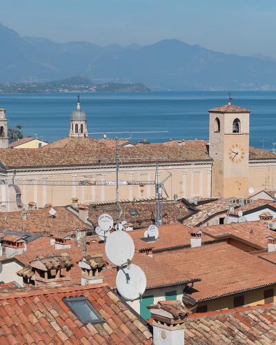 A high-angle view of the terracotta rooftops of Desenzano del Garda, featuring the Duomo di Santa Maria Maddalena with its distinctive bell tower and clock, overlooking Lake Garda with mountains in the background.