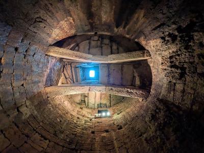 A view looking down into a deep, circular stone well with a spiral staircase winding down its walls and a bright opening at the top.