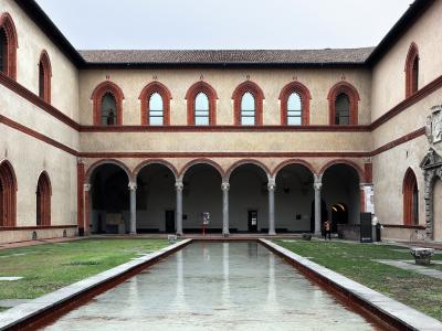 The image shows a rectangular courtyard with a long, narrow reflecting pool in the center, surrounded by grass and flanked by two-story arcades with red-brick arches and columns; signage and a coat of arms are visible on the right wall.