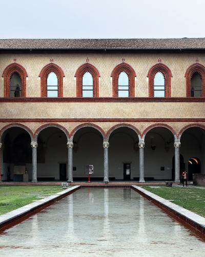 The image shows a rectangular courtyard with a long, narrow reflecting pool in the center, surrounded by grass and flanked by two-story arcades with red-brick arches and columns; signage and a coat of arms are visible on the right wall.