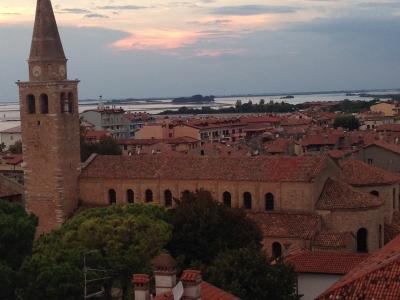 A tall brick bell tower with a pointed spire topped by a statue, adjacent to a long basilica with arched windows and a terracotta roof, overlooking a coastal town with red-tiled roofs and water in the background under a twilight sky.