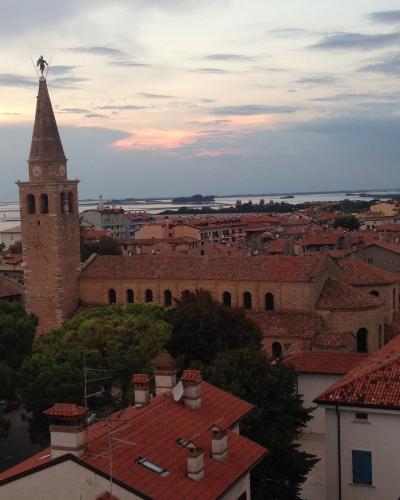 A tall brick bell tower with a pointed spire topped by a statue, adjacent to a long basilica with arched windows and a terracotta roof, overlooking a coastal town with red-tiled roofs and water in the background under a twilight sky.