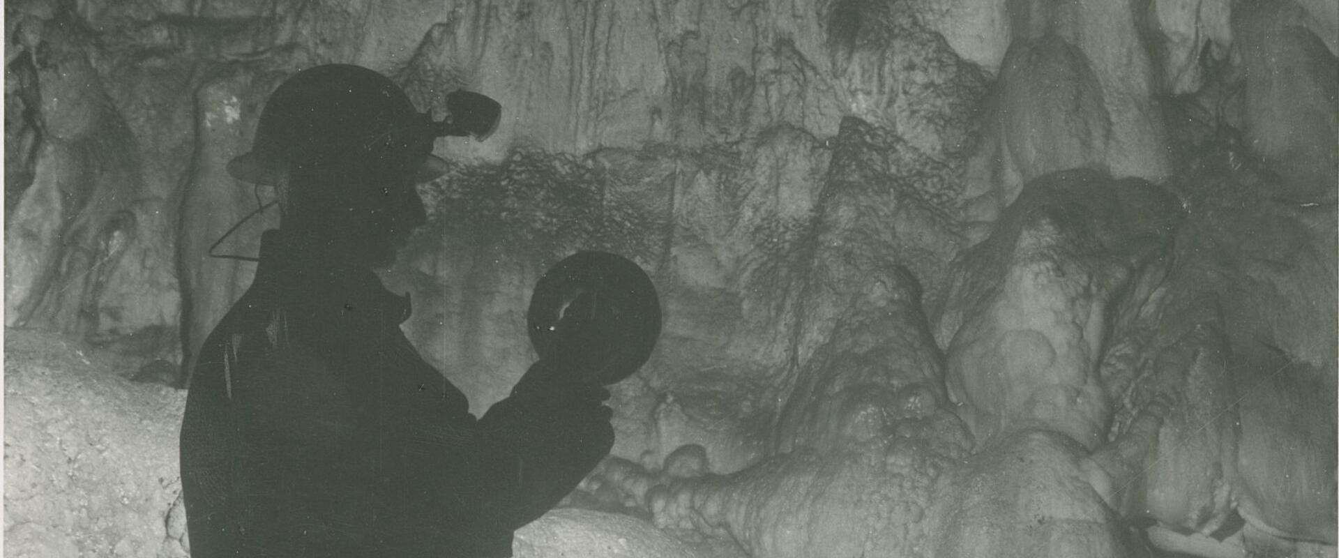A black-and-white photograph showing a silhouetted speleologist wearing a helmet with a headlamp and holding a handheld light, standing inside a cave with prominent stalactites, stalagmites, and flowstone formations; the image includes archival me