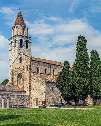 The image shows the Basilica di Santa Maria Assunta in Cielo e Santi Ermagora e Fortunato in Aquileia, Italy, featuring its distinctive Romanesque bell tower, stone basilica structure, adjacent ancient ruins, and a row of tall cypress trees under