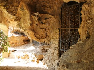 The image shows the interior of a natural rock cave with rough, textured walls, a dirt floor, and a black metal gate installed within an opening on the right side.
