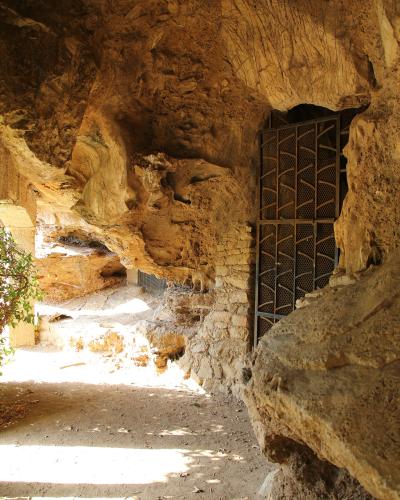 The image shows the interior of a natural rock cave with rough, textured walls, a dirt floor, and a black metal gate installed within an opening on the right side.