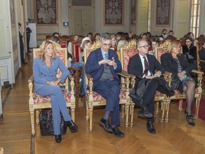 The image shows a formal indoor event with several people seated in ornate, gilded chairs within a historic hall featuring wooden floors and decorative wall panels; the filename identifies the subject as the UK Ambassador to the University of Pavi