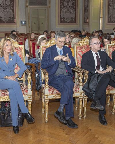 The image shows a formal indoor event with several people seated in ornate, gilded chairs within a historic hall featuring wooden floors and decorative wall panels; the filename identifies the subject as the UK Ambassador to the University of Pavi
