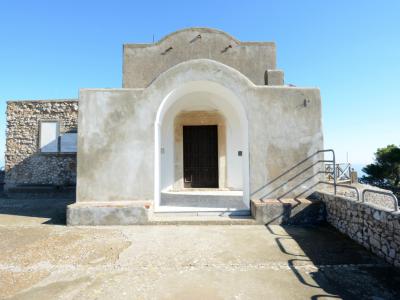 The image shows a small, whitewashed stone chapel with a prominent arched entrance and a simple gabled roof, situated on a paved terrace with a stone wall and sea view in the background; no visible signage or text is present.