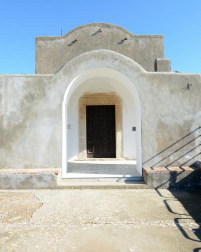 The image shows a small, whitewashed stone chapel with a prominent arched entrance and a simple gabled roof, situated on a paved terrace with a stone wall and sea view in the background; no visible signage or text is present.