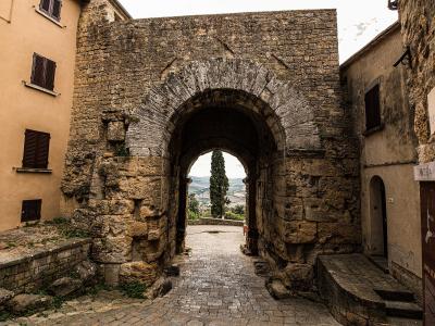 A view from inside looking out through the large stone archway of Porta all'Arco in Volterra, Italy, with a sign on the right reading "VIA PORTA ALL'ARCO" and another sign below it mentioning "PORTA ALL'ARCO".