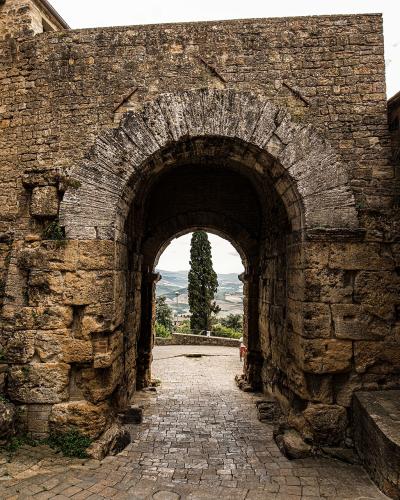 A view from inside looking out through the large stone archway of Porta all'Arco in Volterra, Italy, with a sign on the right reading "VIA PORTA ALL'ARCO" and another sign below it mentioning "PORTA ALL'ARCO".