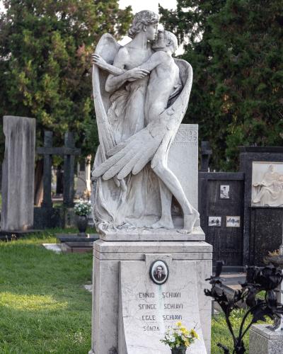 A white marble funerary monument featuring a sculpted angel embracing a deceased figure, with the surname "SCHIavi" inscribed on the base, set within a grassy cemetery with other tombs and trees in the background.
