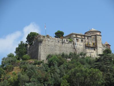 A stone fortress with crenellated walls, a round tower, and yellow-shuttered windows sits atop a green hill under a blue sky, with an Italian flag flying on a pole near the top.