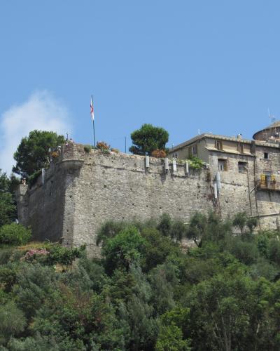 A stone fortress with crenellated walls, a round tower, and yellow-shuttered windows sits atop a green hill under a blue sky, with an Italian flag flying on a pole near the top.