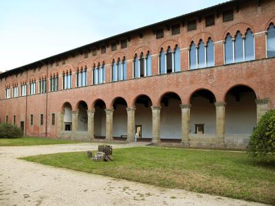 The image shows the exterior of a large, two-story red-brick building with a ground-floor loggia featuring stone columns and arches, and an upper floor with a series of tall, arched windows; the setting is a grassy courtyard with a gravel path.