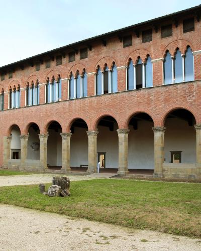 The image shows the exterior of a large, two-story red-brick building with a ground-floor loggia featuring stone columns and arches, and an upper floor with a series of tall, arched windows; the setting is a grassy courtyard with a gravel path.