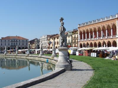 The image shows a large, oval-shaped public square featuring a central grassy island surrounded by a canal, lined with statues on pedestals, with market stalls set up under white canopies along the perimeter and a distinctive building with a long