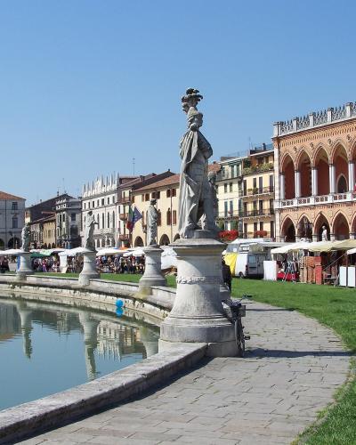The image shows a large, oval-shaped public square featuring a central grassy island surrounded by a canal, lined with statues on pedestals, with market stalls set up under white canopies along the perimeter and a distinctive building with a long