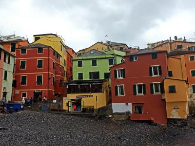 A pebbled beach in the foreground with a cluster of colorful, multi-story buildings rising behind it, including a yellow building with signage reading "Colonia Genovese di Mare" and a red building with green shutters; people are visible near the b