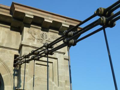 A close-up view of the stone anchor tower and thick suspension cables of a chain bridge, featuring decorative stone carvings and large metal cable clamps against a blue sky.