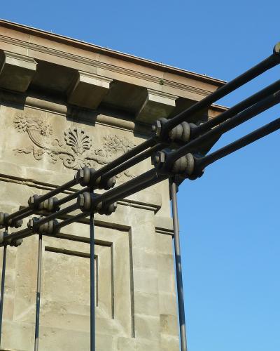 A close-up view of the stone anchor tower and thick suspension cables of a chain bridge, featuring decorative stone carvings and large metal cable clamps against a blue sky.