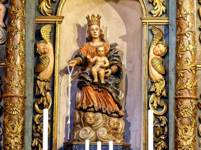 A close-up interior shot of an ornate, gilded Baroque-style altarpiece featuring a central statue of the Virgin Mary and Child surrounded by cherubs, flanked by twisted columns and candles.