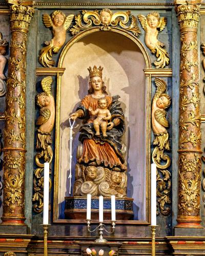 A close-up interior shot of an ornate, gilded Baroque-style altarpiece featuring a central statue of the Virgin Mary and Child surrounded by cherubs, flanked by twisted columns and candles.