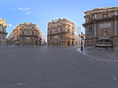 A 360-degree panoramic photograph of an octagonal city square featuring four ornate, curved corner buildings with statues and fountains, under a blue sky with some construction scaffolding visible.